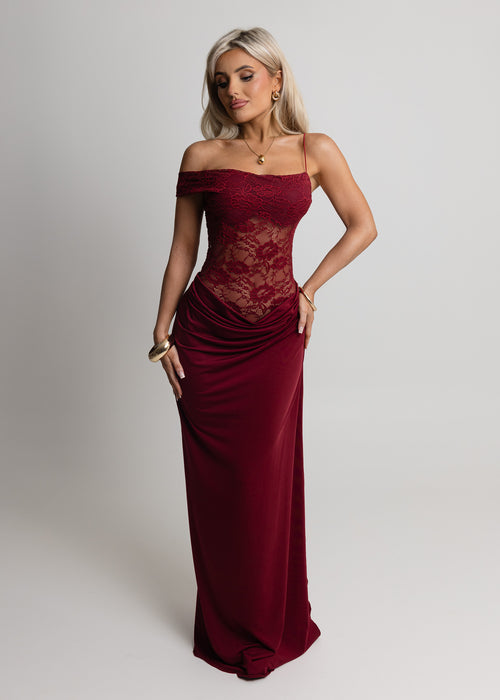 Woman standing against light backdrop in a studio, wearing a burgundy lace maxi dress with Lycra fitting maxi skirt, paired with gold jewellery and posed with one hand on hip.