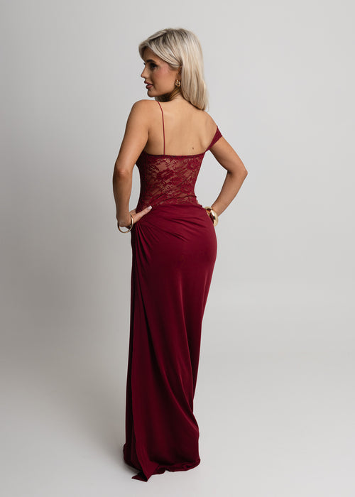 Woman standing against light backdrop in a studio, wearing a burgundy lace maxi dress with Lycra fitting maxi skirt, paired with gold jewellery and posed with one hand on hip.