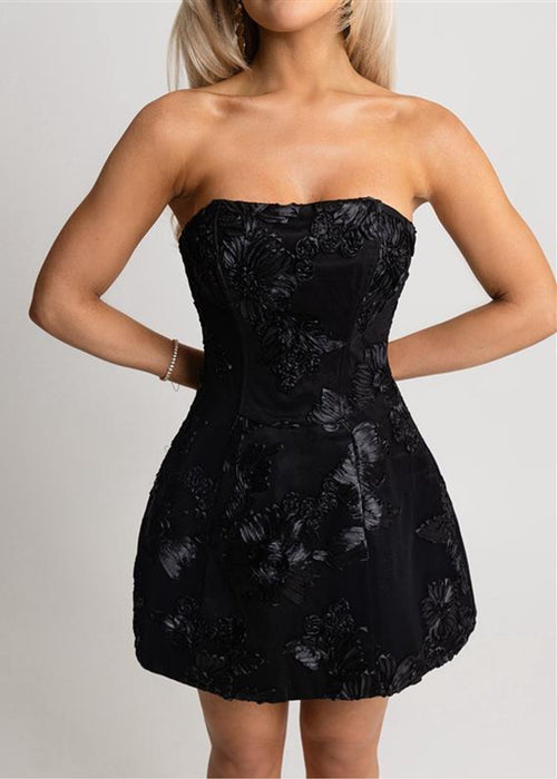 Woman standing against a light background wearing an embroidered black strapless mini dress with A-line silhouette and paired with gold high heels, posing with one hand on hip.