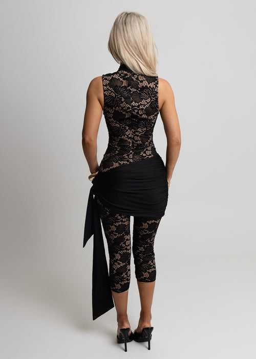Woman standing against a light backdrop in a studio wearing a black lace jumpsuit with keyhole cut out neckline, jersey sarong style detailing across waist and capri length pants, paired with gold jewellery, posed with hands resting on thighs.
