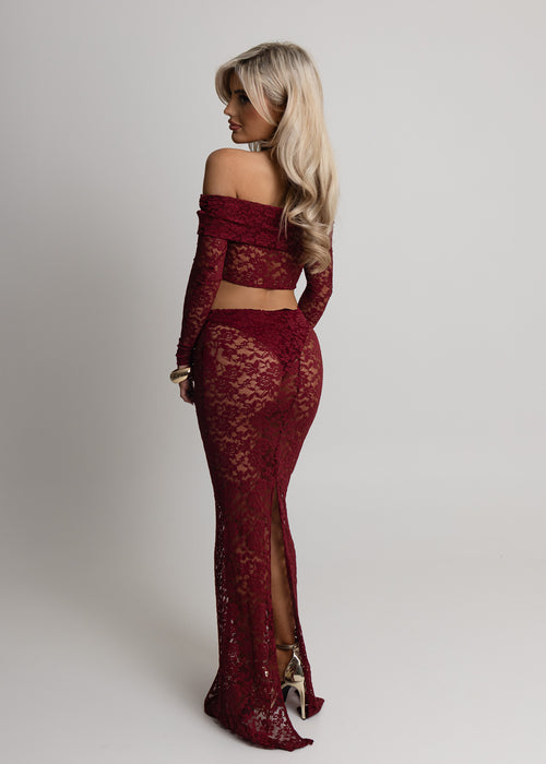Woman standing against a light  backdrop in a studio wearing a burgundy lace two piece set with long sleeve sheer lace crop top and matching lace maxi skirt, item comes with undergarments. Paired with gold jewellery and posed with both hands relaxed on thighs. 