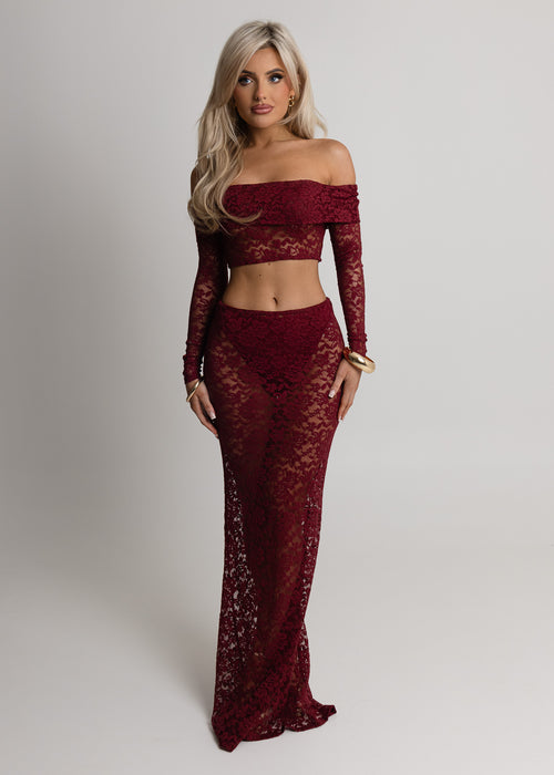Woman standing against a light  backdrop in a studio wearing a burgundy lace two piece set with long sleeve sheer lace crop top and matching lace maxi skirt, item comes with undergarments. Paired with gold jewellery and posed with both hands relaxed on thighs. 