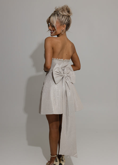Women's champagne glitter mini dress with large detachable bow on the back of mini dress. Pearl and diamante trim embellishments with a strapless fit. Model is 5ft2' and wears a size XS 6.